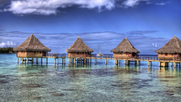 A row of bungalow houses stands on stilts over clear blue water under a partly cloudy sky, 4K Desktop Wallpaper