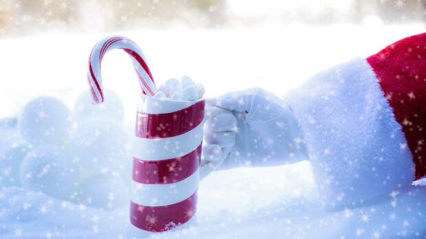 A candy cane held in a hand wearing a white glove with a striped mug filled with marshmallows in the snow