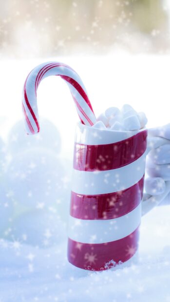 A festive candy cane leaning inside a striped ceramic mug filled with marshmallows in the snow