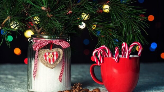 A festive arrangement of candy cane sticks in a red mug next to a decorated jar with pine branches and a heart ornament