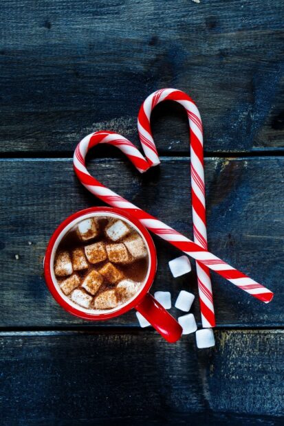 Two candy canes forming a heart shape next to a red cup of hot chocolate with marshmallows on a wooden table