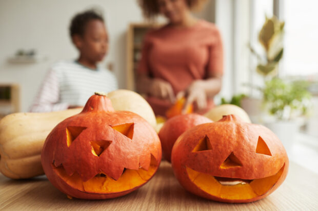 Two carved pumpkins sit on a table with a child and adult in the background preparing more pumpkins, HD Desktop Wallpaper