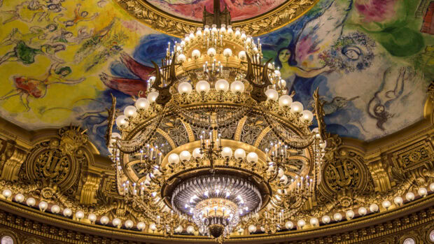 Gold chandelier with elaborate design hanging from a painted ceiling mural in a grand hall