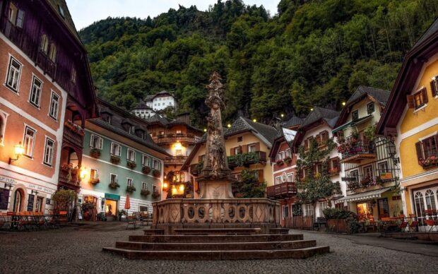 The charming village square in Austria is surrounded by colorful houses and a detailed stone monument, HD Desktop Wallpaper