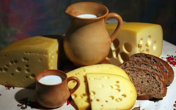A variety of cheese pieces with holes displayed alongside sliced bread and ceramic jugs of milk on a tablecloth