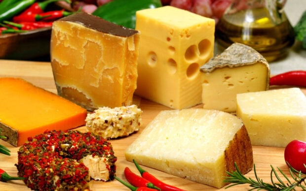 Various cheese types arranged on wooden board with herbs and spices surrounding