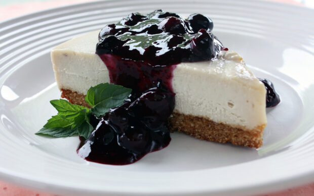 A slice of cheese cake topped with blueberry sauce and garnished with mint leaves on a white plate