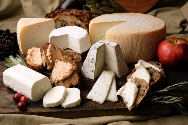 A variety of cheese arranged on a wooden board with bread and an apple nearby