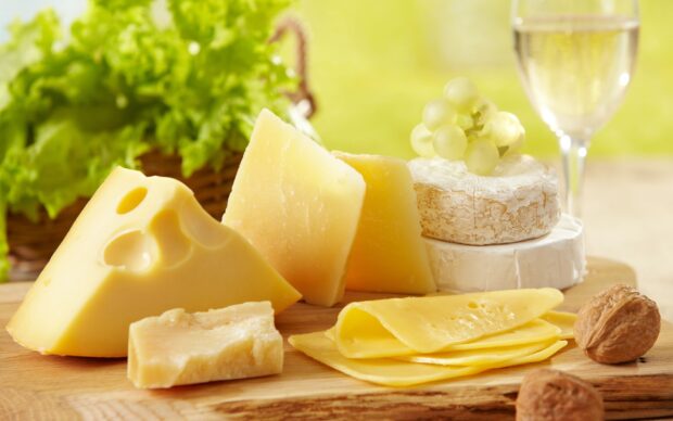 A variety of cheese including slices and wedges arranged on a wooden board with grapes and walnuts in the background