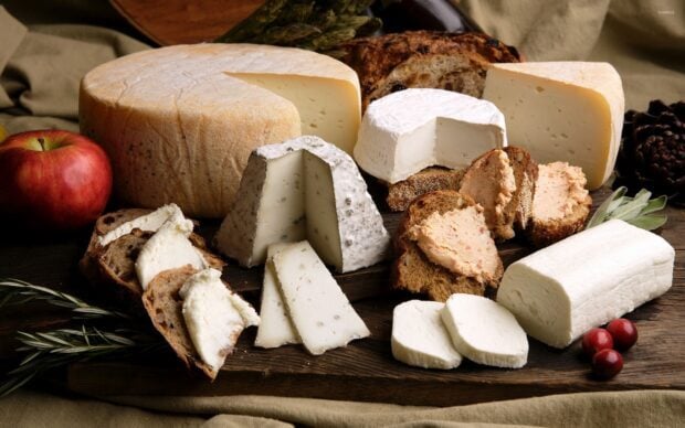 A variety of cheese pieces arranged on a wooden board with bread and an apple nearby