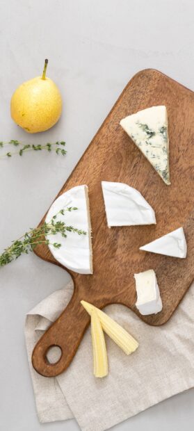 A wooden board with different types of cheese and fresh herbs arranged next to a yellow pear and baby corn