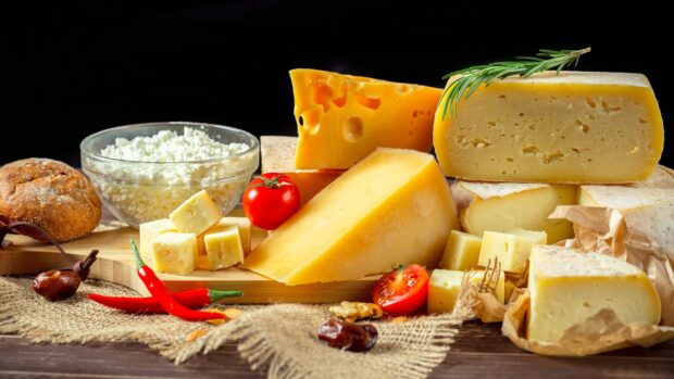A variety of cheese pieces including cottage cheese and cheddar on a wooden board with tomatoes and chili peppers