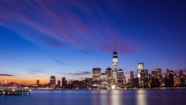 A city skylines view at dusk with illuminated skyscrapers and a calm waterfront scene