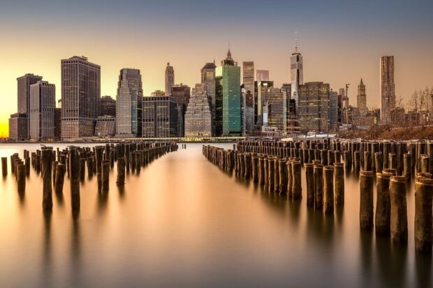 Wooden pillars in water leading to city skylines at sunset with calm reflections