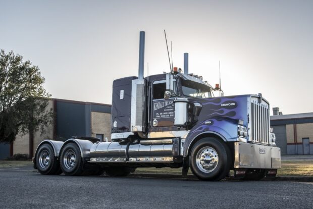 A classic Kenworth truck with purple flames is parked near industrial buildings under a clear sky, HD Desktop Wallpaper