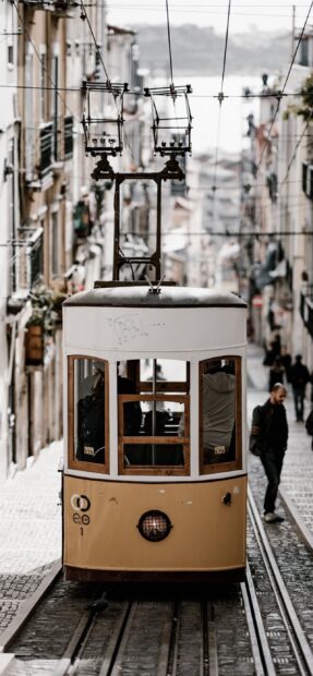 A classic tram moving through narrow streets in Lisbon Portugal with pedestrians around, 2K Mobile Wallpaper