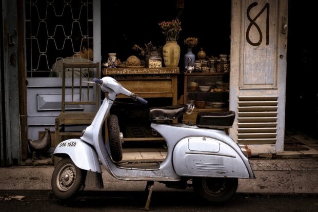A classic blue scooter is parked outside a rustic shop with vintage decor and a person sitting inside, HD Desktop Wallpaper