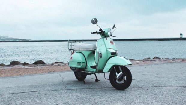 A mint green scooter is parked on the road by the waterfront under a cloudy sky with calm water in the background