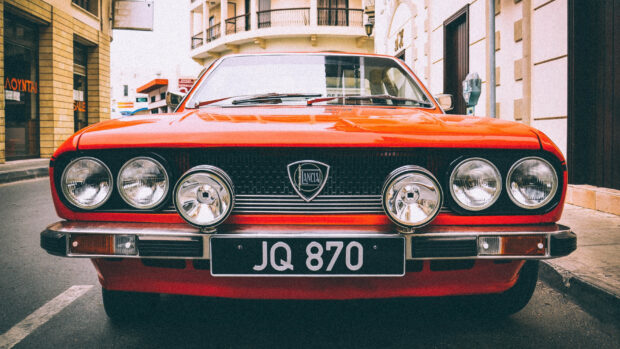 A classic red Lancia car is parked on a street with buildings around, 2K Desktop Wallpaper