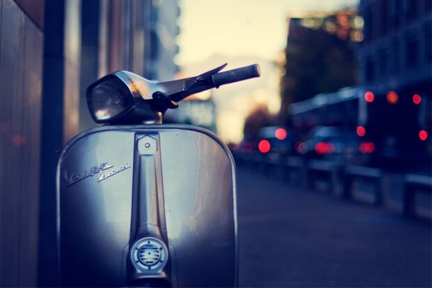 A classic scooter is parked on a city street with blurred lights in the background during dusk, HD Desktop Wallpaper