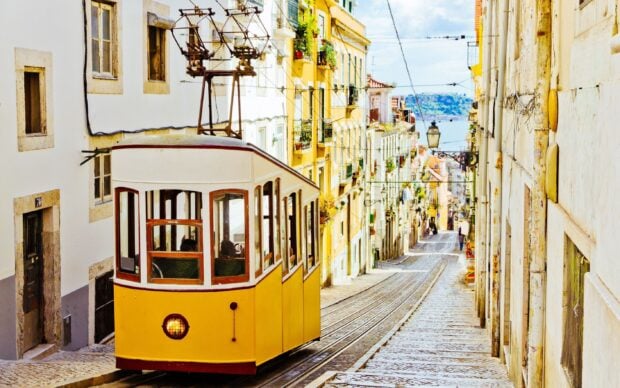 A classic yellow tram climbs a steep street lined with buildings in Lisbon Portugal, 2K Desktop Wallpaper