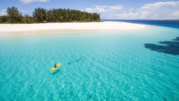 A person is kayaking in clear turquoise water near a sandy beach in Zanzibar, 2K Desktop Wallpaper