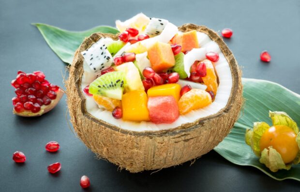 Fresh coconut bowl filled with tropical fruit pieces on a table with green leaves and pomegranate seeds