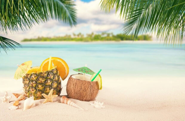 A tropical coconut drink with a small umbrella and straw on a sandy beach with palm leaves overhead