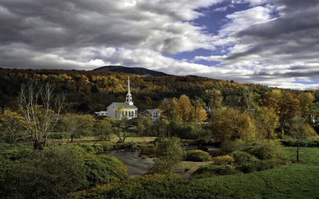A peaceful Vermont scene shows a white church among autumn trees and hills under a partly cloudy sky, HD Desktop Wallpaper