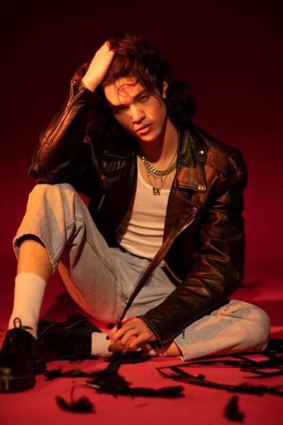 Conan Gray sits on a red surface wearing a leather jacket and chains, 2K Mobile Wallpaper