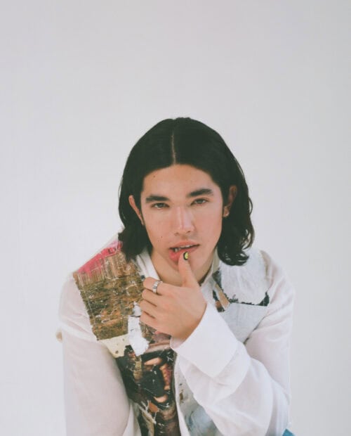 A young man with dark hair wearing a white graphic shirt is posing thoughtfully in this Conan Gray wallpapers, HD Mobile Wallpaper