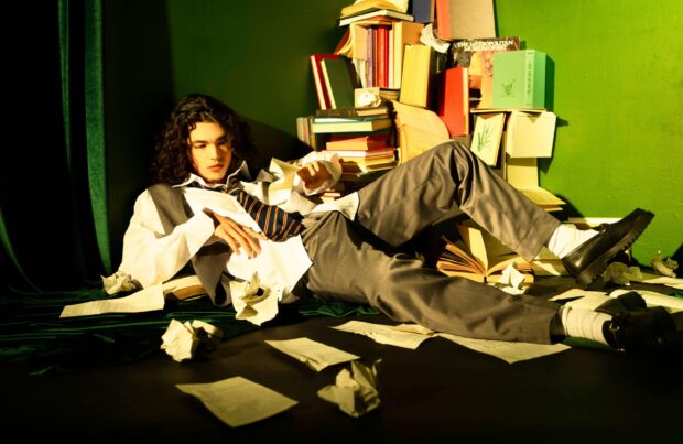A person in a tie and vest lies among books and papers in a green room featuring Conan Gray wallpapers, 2K Desktop Wallpaper