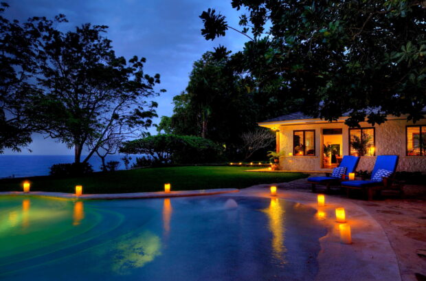 A tropical bungalow is illuminated warmly by candlelight beside a pool at twilight, 2K Desktop Wallpaper