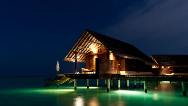 A bungalow illuminated by warm lights stands over glowing blue water at night, HD Desktop Wallpaper