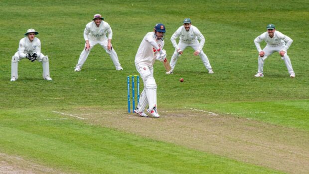 A cricket player batting on the pitch with fielders positioned behind him on the grassy field