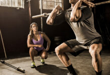 Female fitness trainer coaching man lifting barbell in gym.