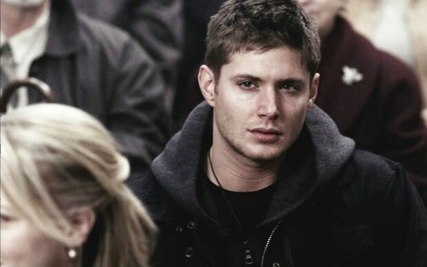 Dean Winchester wearing a black jacket and hoodie looking intently in a crowded room