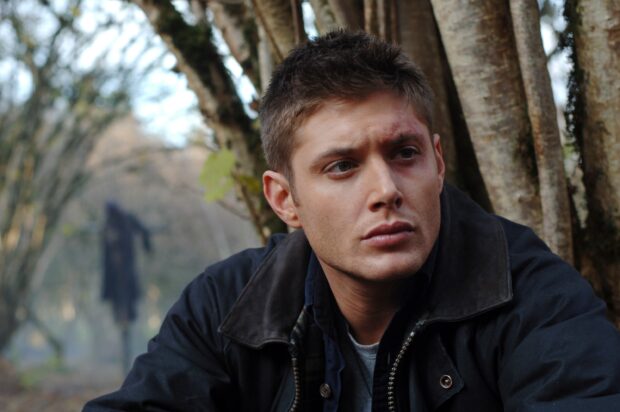 Dean Winchester with a bruised face sitting outdoors near a tree in a forest setting