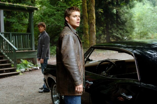 Dean Winchester stands by a car in the forest with another man in the background
