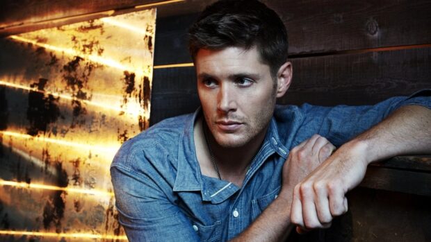 Dean Winchester wearing a blue shirt leaning against a wooden wall in a moody setting