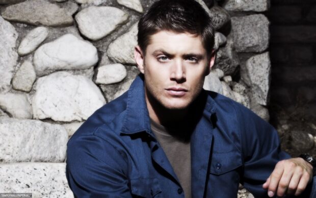 Dean Winchester in a blue shirt sitting against a stone wall looking serious