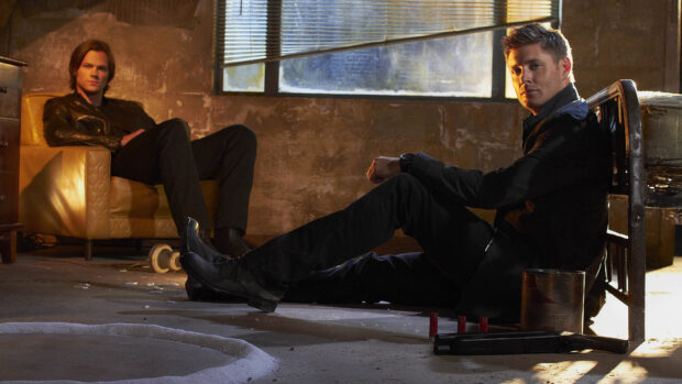 Dean Winchester sitting on the floor in a dark room with another man in the background