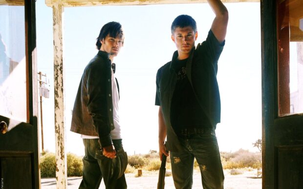 Dean Winchester standing with his brother in a desert scene holding weapons