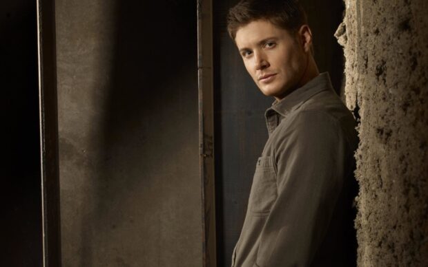 Dean Winchester standing against a textured wall in a casual shirt looking at the camera