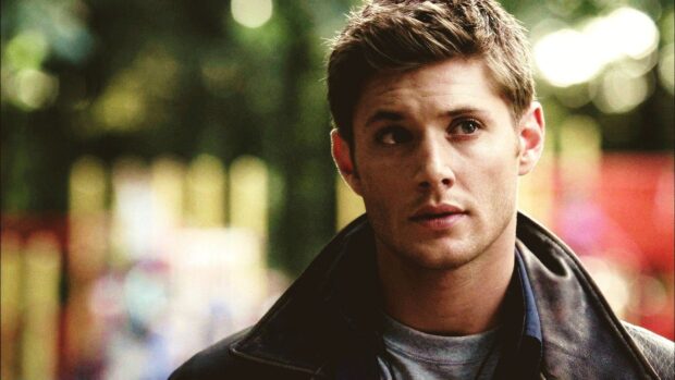 Dean Winchester with a serious expression wearing a dark jacket outdoors in a blurred background