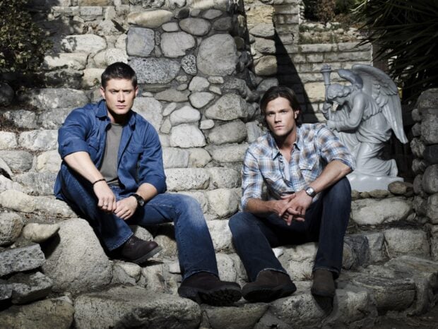 Dean Winchester sitting on stone steps with a man in casual clothes in a rustic setting