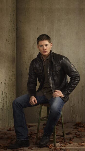 Dean Winchester sitting on a stool wearing a leather jacket and jeans in a rustic setting