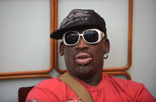 Dennis Rodman wearing sunglasses and a cap with facial piercings and a red shirt indoors