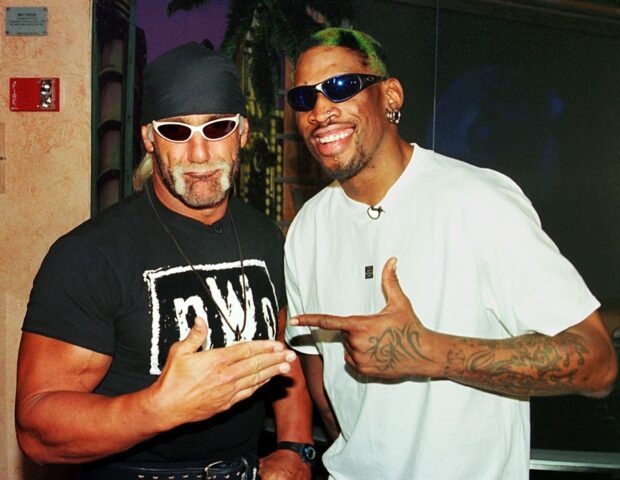 Dennis Rodman smiling with a tattooed arm and colorful hair standing next to a muscular man wearing a black bandana and nWo shirt