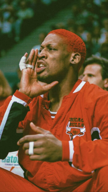 Dennis Rodman with red hair wearing a Chicago Bulls jacket shouting on the bench